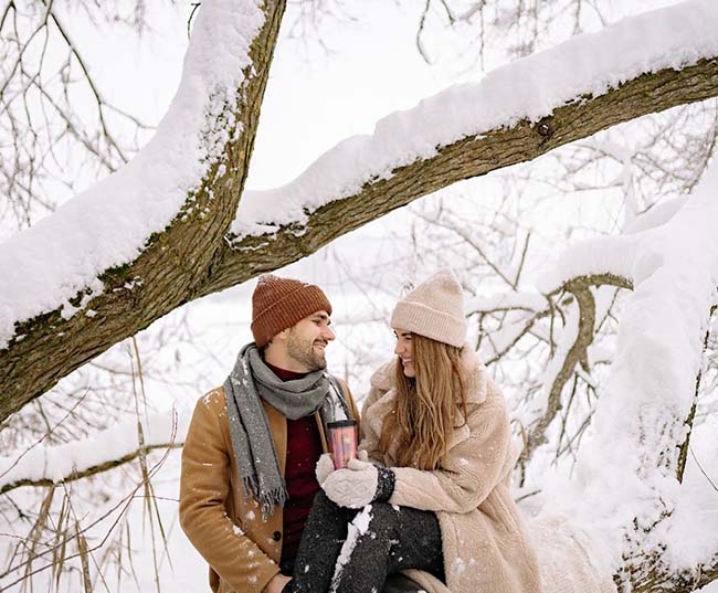 A lovely couple in winter clothes looking at each other under the snow covered tree branch