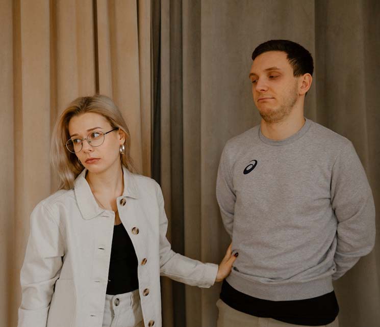 A man in gray sweater standing beside a woman in beige blazer feeling disappointed