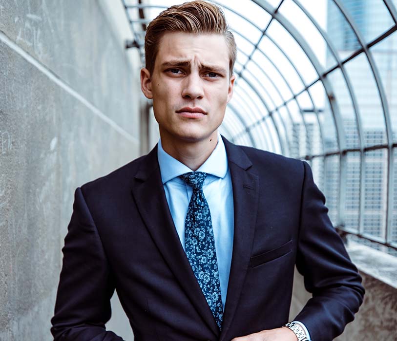 A man wearing black and teil dress suit standing near grey wall