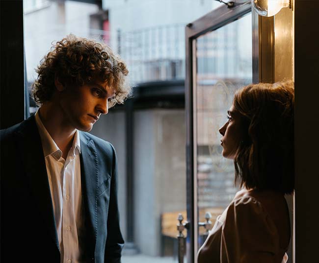 A woman in a brown coat and a man in a black coat stand facing each other looking at each other.